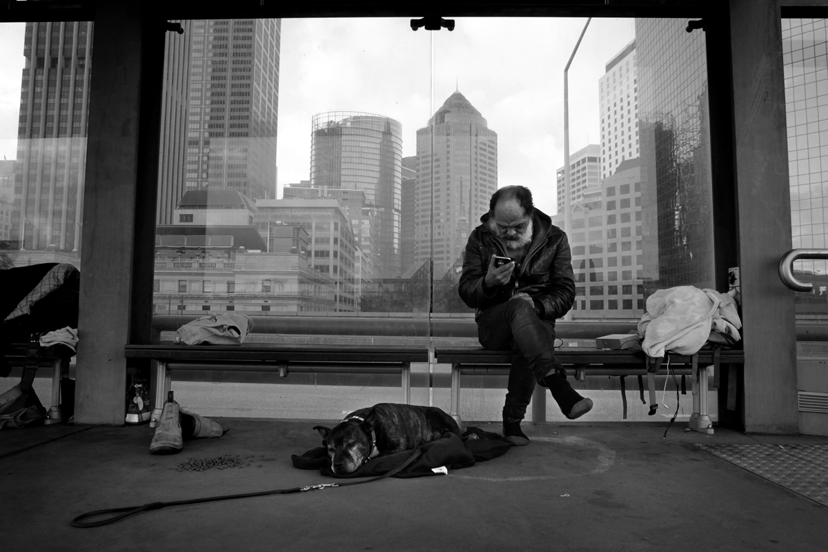Lanz Priestly sits on his bench, looking at his phone, with the city's skyscrapers in the background.