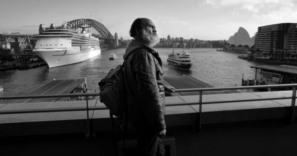 Lanz Priestly stands on the railing of the Cahill Expressway. In the background, to his left, you can see the Harbour Bridge and a docked cruise ship. The the right are the sails of the Opera House.