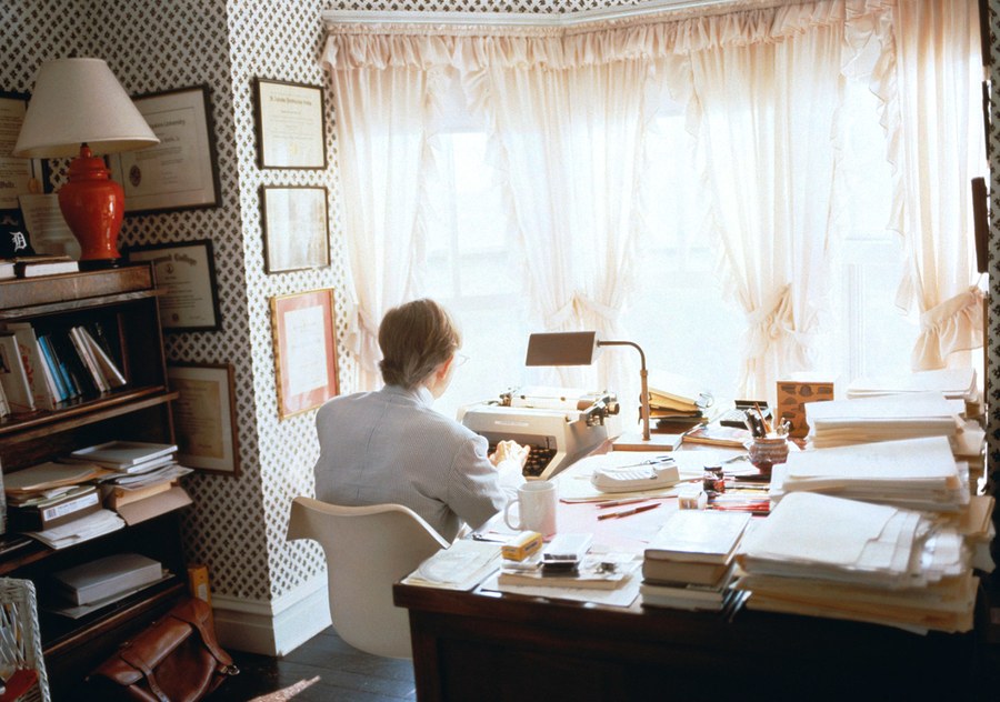 Wolfe at his desk in a white suit, facing away from the camera, using a type-writer