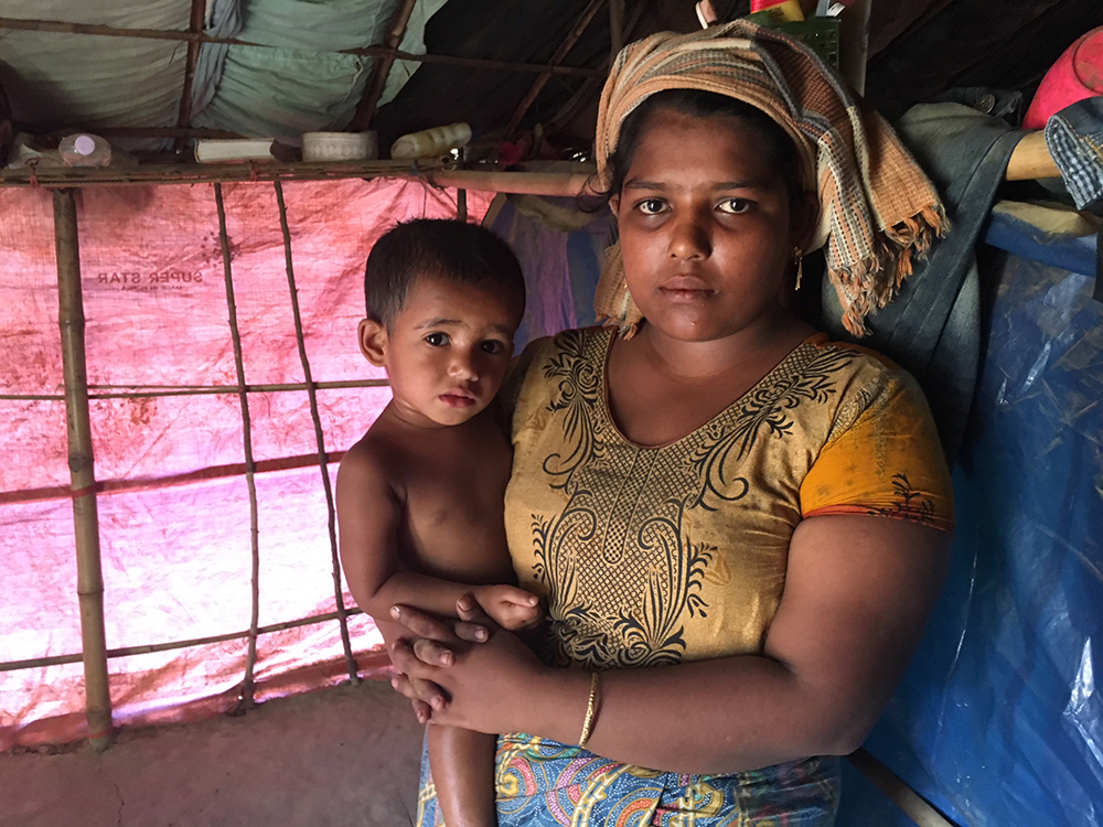 A Rohingya mother holds her child in a makeshift tent made of bamboo and red sheet plastic